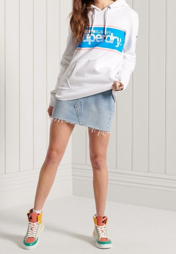 Personne portant un sweat à capuche blanc avec le logo bleu Superdry, une jupe en denim claire effilochée et des baskets montantes colorées, debout dans une pièce lumineuse.