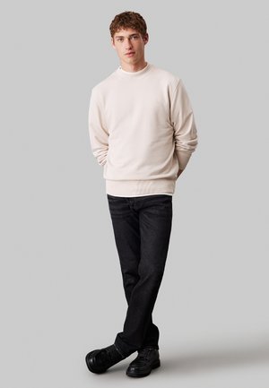 Felpa beige chiaro con scollo e polsini a costine. Jeans in denim scuro e scarpe nere. Il modello è in piedi con le mani in tasca.