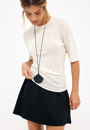 Femme portant un haut blanc à manches courtes et une jupe noire, regardant un long collier avec un pendentif en forme de coquillage noir.