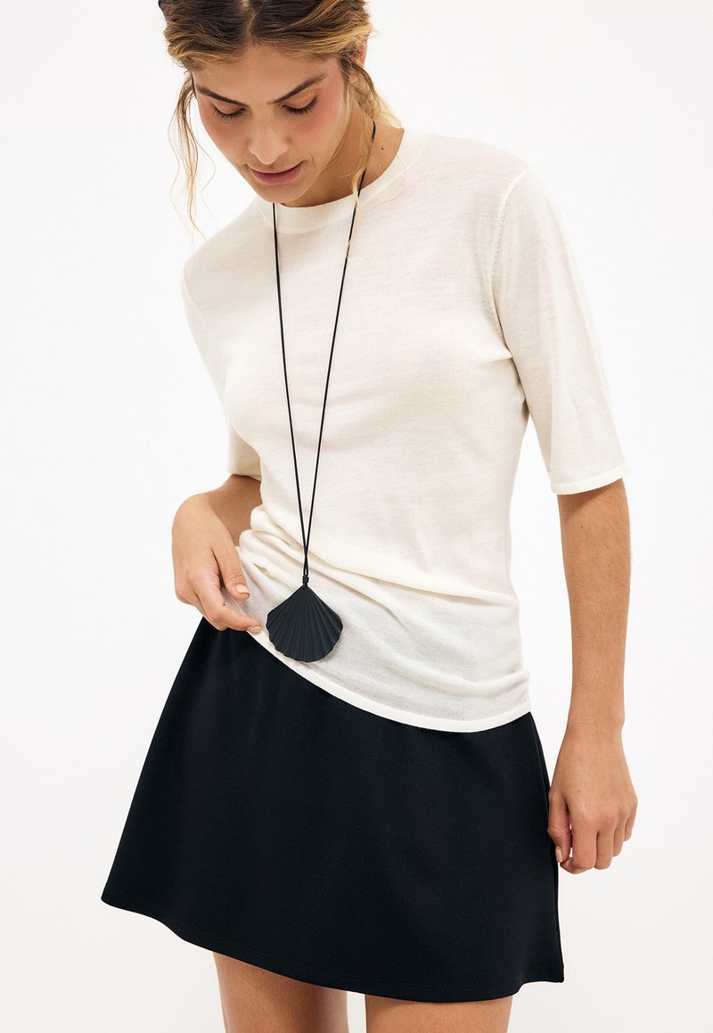 Kvinde iført en hvid top med korte ærmer og sort nederdel, der ser ned på en lang halskæde med en sort, skaldyrsformet vedhæng.