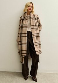 Blond kvinna i beige rutiga långrock, ljus tröja, svarta vida byxor och spetsiga klackskor som står mot en enfärgad vägg.