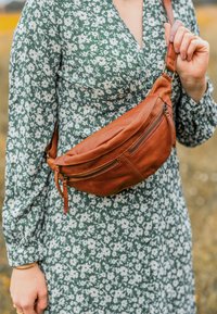Marsupio in pelle marrone con una consistenza morbida, caratterizzato da due scomparti con zip e una tracolla regolabile, indossato sopra un vestito con fantasia floreale.