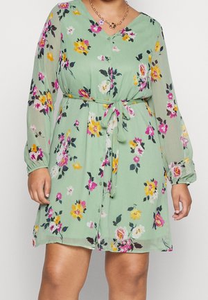 Frau trägt ein hellgrünes Kleid mit floralem Muster, langen transparenten Ärmeln und einem Taillengürtel, kombiniert mit zwei goldenen Kettenhalsketten.