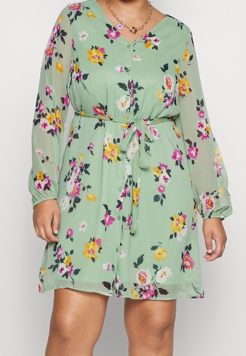 Femme portant une robe à fleurs vert clair avec de longues manches transparentes et une ceinture nouée à la taille, accessoirisée de deux colliers chaînes dorés.