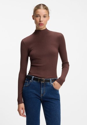 Vrouw met lichtbruin haar in een staart, draagt een aansluitende bruine geribbelde longsleeve en blauwe jeans met een zwarte riem, staand met één hand in de zak.