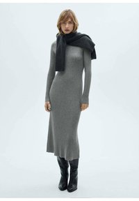 Robe en tricot côtelé grise avec des manches longues, design à mi-genoux, associée à une écharpe sombre. Le mannequin porte des bottes noires à hauteur de genou.