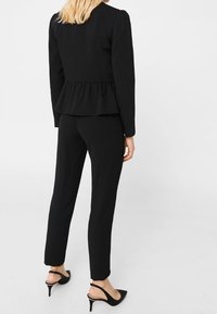 Zwarte op maat gemaakte pak met een peplumdetail in de taille. De top heeft lange mouwen, gecombineerd met slim-fit broek en puntige zwarte hakken.