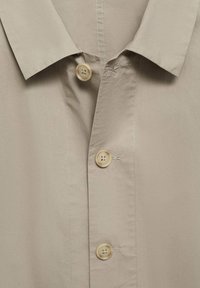 Jasno beżowa koszula z wyraźnym kołnierzykiem, z trzema jasnobrązowymi guzikami i gładką strukturą materiału. Widoczne przeszycia wzdłuż kołnierzyka.