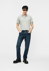 Άνδρας που φοράει ανοιχτό γκρι πλεκτή μπλούζα πόλο, σκούρα μπλε τζιν, μαύρα χοντρά παπούτσια και μαύρα γυαλιά ηλίου, όρθιος με τα χέρια στις τσέπες.