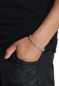 Bracciale in argento con anelli intrecciati, indossato su un polso, con una superficie liscia, su sfondo di camicia e pantaloni neri.