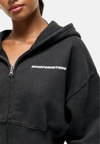 Felpa nera con zip realizzata in materiale morbido, con un logo bianco "GOODFORNOTHING" sul lato sinistro. Include un cappuccio foderato e polsini a costine.