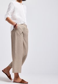 Person klädd i beige randiga cropped byxor, vit långärmad skjorta med knappar, bruna sandaler och ett brunt armband, som står med händerna i fickorna.