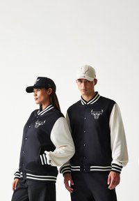 Μαύρα και κρεμ varsity μπουφάν με λογότυπο των Chicago Bulls, με ριμπ μανσέτες, γιακά και κουμπιά. Δύο ταιριαστά καπέλα: μαύρο και κρεμ.
