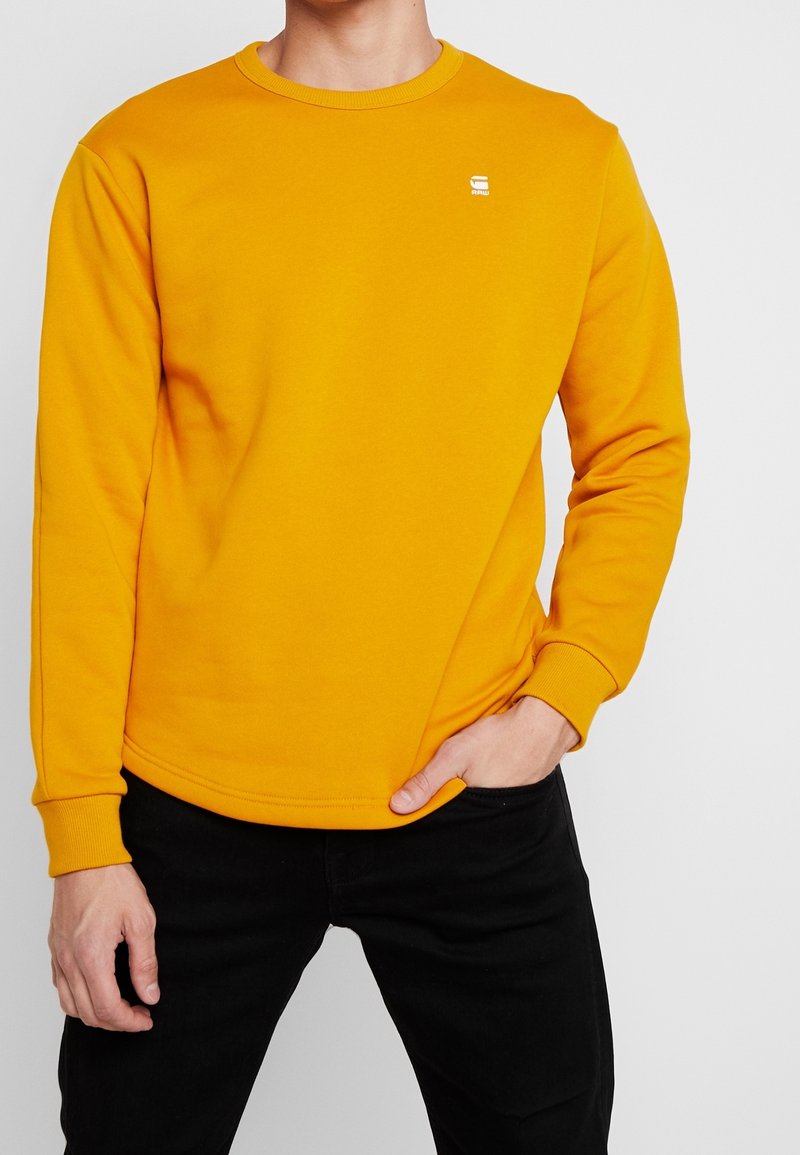 Sweat-shirt jaune avec un col rond, des poignets côtelés et un ourlet côtelé. Dispose d'un petit logo blanc sur la poitrine. Porté avec un pantalon noir.