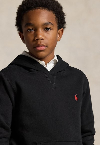Chlapec s krátkymi kučeravými vlasmi, ktorý má na sebe čiernu mikinu s kapucňou cez bielu košeľu s golierom na neutrálnom pozadí.