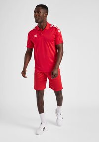Rode polo shirt en shorts set, met witte schouderpatronen en logo. Draagt met witte schoenen en sokken, wat een casual sportieve uitstraling laat zien.