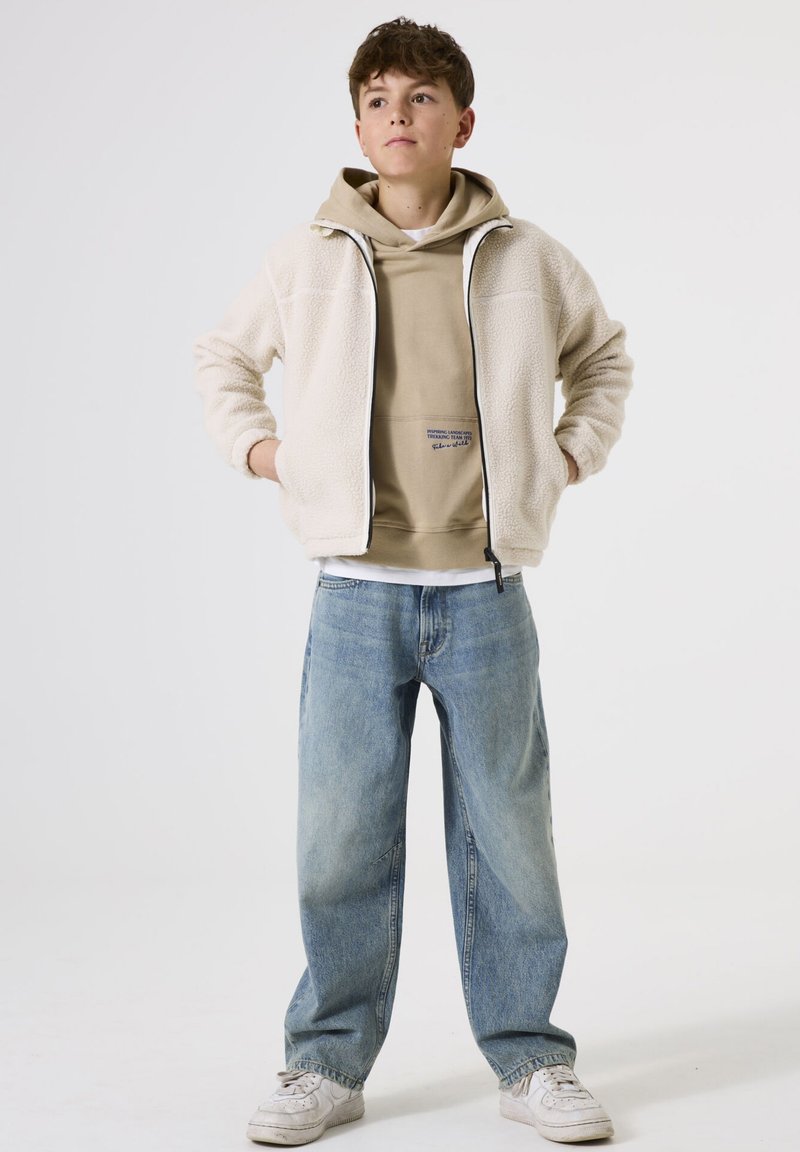 Chaqueta blanca y peluda con capucha sobre una sudadera beige, vaqueros holgados de color azul claro y zapatillas blancas, en un fondo liso.