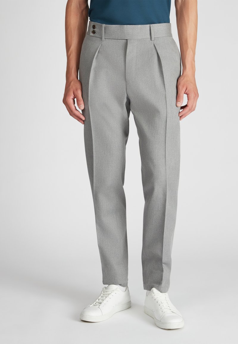 Man in een lichte, grijze, op maat gemaakte broek met voorplooien en witte sneakers, staande tegen een effen achtergrond.