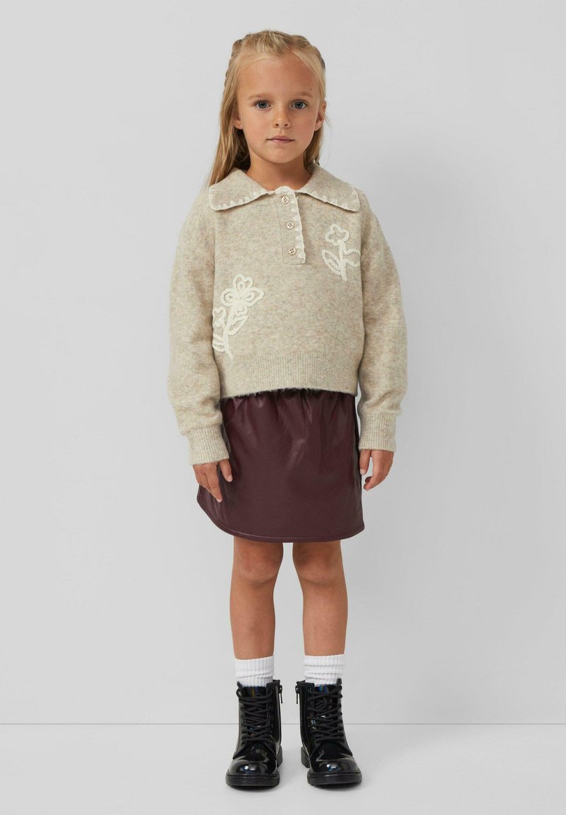 Beige gebreide trui met bloemengaren, gecombineerd met een bordeaux neplederen rok. Zwarte veterschoenen en witte kniekousen.
