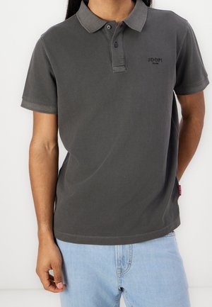 Homme portant un polo à manches courtes gris foncé avec des boutons et un jean bleu clair, debout devant un fond blanc uni.