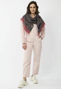 Salopette en coton rose avec poches avant, accompagnée d'une écharpe à motifs noirs et rouges, portée avec des baskets blanches sur un fond uni.