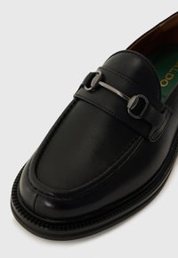 Schwarzer Lederloafer mit glatter Oberfläche, ausgestattet mit einem metallischen Akzentriemen und detaillierter Naht entlang der Sohle. Klassisches Design.