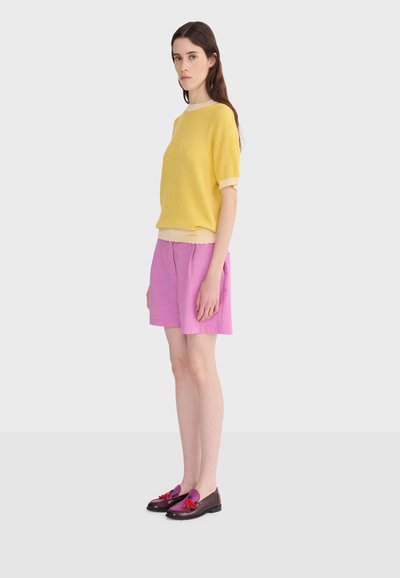 Jeune femme debout, portant un haut jaune à manches courtes, un short rose clair et des mocassins foncés avec des pompons violets et rouges, sur un fond uni.