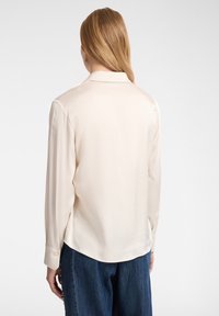Blusa di seta color beige chiaro con colletto, maniche lunghe e polsini con bottoni. Tessuto liscio, orlo leggermente curvato, indossata con pantaloni in denim larghi.