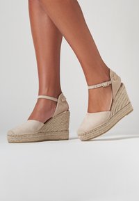 Beige ruskind kilehæle med fletet jutesål, justerbar ankelrem og rundet tå, der viser en struktureret overdel og et let design.