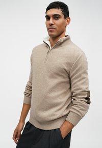 Bežs adīts džemperis ar ceturtdaļas rāvējslēdzēja apkakli, strukturētu audumu, ribotām piedurknēm un smalku logo detaļu uz krūšu. Saskaņots ar tumšām biksēm.