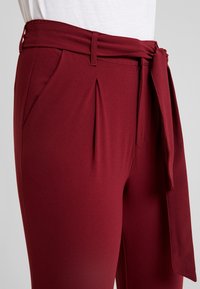 Bordeauxkleurige broek van zachte stof, voorzien van een gestrikt tailleband, voorplooien en zijzakken voor extra detail en functionaliteit.