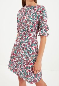 Blumenkleid mit weißem Hintergrund, versehen mit lebendigen roten, rosa und grünen Mustern. Rundhalsausschnitt und geraffte, gep puffte Ärmel.