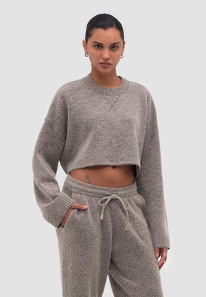 Vrouw met een grijze cropped trui en bijpassende sweatpants met trekkoord, met handen in de zakken, staand tegen een effen achtergrond.