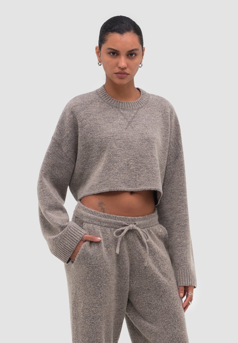 Femme portant un sweat gris court et un pantalon de survêtement assorti à cordon, les mains dans les poches, debout devant un fond uni.