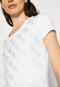 Hvid kortærmet t-shirt med et struktureret mønster af skinnende sølvdetaljer hen over fronten. V-udskæring, blødt materiale.