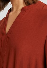 Vaate, jonka väri on ruosteinen ja pehmeä tekstuuri, V-pääntie. Sisältää hienovaraisia ompeleita ja rentoa istuvuutta hartioiden ympärillä.