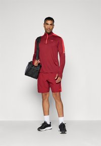 Mand iført rød træningstop og shorts, sorte sneakers med hvide såler, hvide sokker, holder en sort sportstaske med skulderrem.