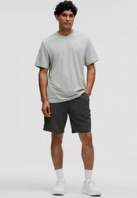 Lys grøn bomulds t-shirt parret med mørkegrå shorts. Modellen står med hænderne i lommerne, hvide træningssko og sokker synlige.