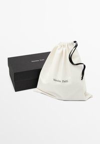 Borsa di cotone con coulisse color crema con logo "Massimo Dutti", accanto a una scatola rettangolare nera, entrambe disposte su uno sfondo bianco.