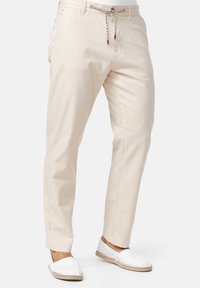 Beige Baumwollhosen mit schmalem Schnitt, ausgestattet mit einem Kordelzugbund und Seitentaschen. Kombiniert mit weißen Slippers.