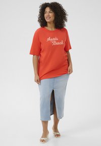 T-shirt rouge avec le texte blanc "Après Beach", à manches courtes, associé à une jupe en denim clair avec une fente à l'avant, et des sandales à talons blancs.