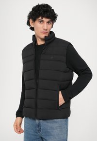 Chaleco acolchado negro con cuello alto, cierre de cremallera y bolsillos laterales, usado sobre una camiseta de manga larga negra, exhibido sobre un fondo claro.