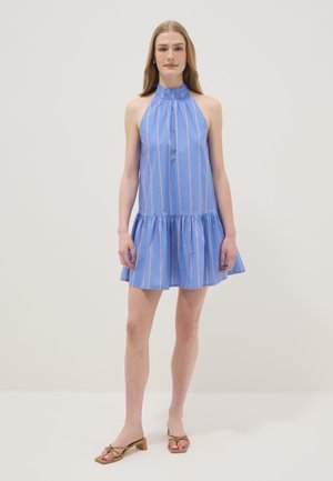 Jonge vrouw staand, gekleed in een mouwloze blauwe halterjurk met verticale strepen en gerimpelde zoom, gecombineerd met beige sandalen met lage hak.