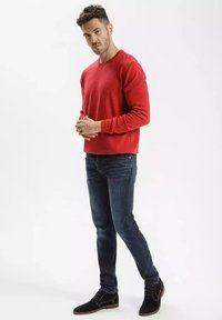 Czerwony sweter z okrągłym dekoltem wykonany z tkaniny, zestawiony z ciemnoniebieskimi dżinsami. Obuwie to czarne zamszowe buty z sznurowadłami i kontrastującymi czerwonymi akcentami.