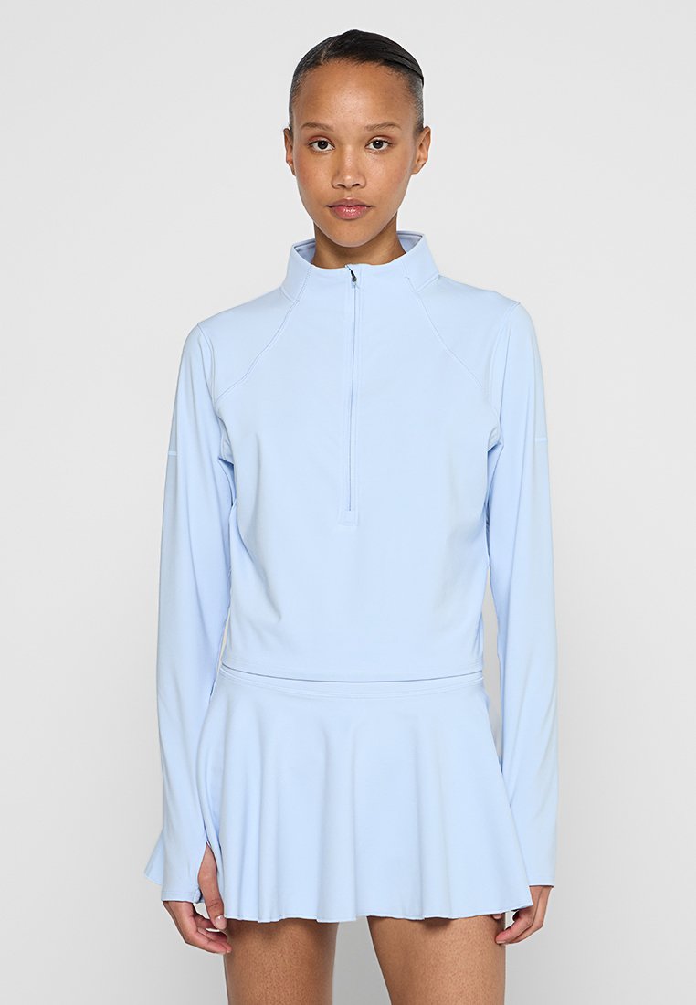 Haut de sport bleu clair à zip quart avec des manches longues, un corsage ajusté et une jupe évasée. Matériau lisse et extensible, design minimaliste.