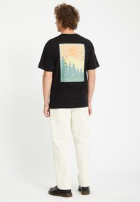 Sort kortærmet T-shirt med et farverigt bjerg- og skovgrafik på bagsiden, med "Volcom" tekst nederst. Set med lyse bukser.
