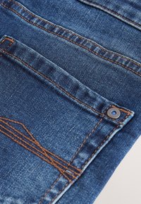 Tessuto di denim blu con cuciture arancioni dettagliate che evidenziano una tasca posteriore e un rivetto su un paio di jeans.