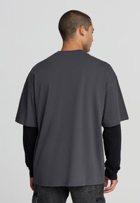 T-shirt grigio a maniche corte, vestibilità comoda, realizzato in morbido cotone, con scollo rotondo e orli delle maniche cuciti. Indossato su una camicia a maniche lunghe nera.