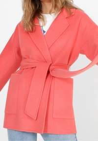 Abrigo mezcla de lana color coral con cinturón, solapas con muesca, dos bolsillos delanteros y ajuste holgado. Textura suave y diseño de longitud media.