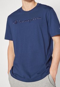 Tmavě modré bavlněné tričko s krátkými rukávy, na hrudi s vyšitým logem "Champion" v tmavě modré barvě. Hladká textura a pravidelný střih.